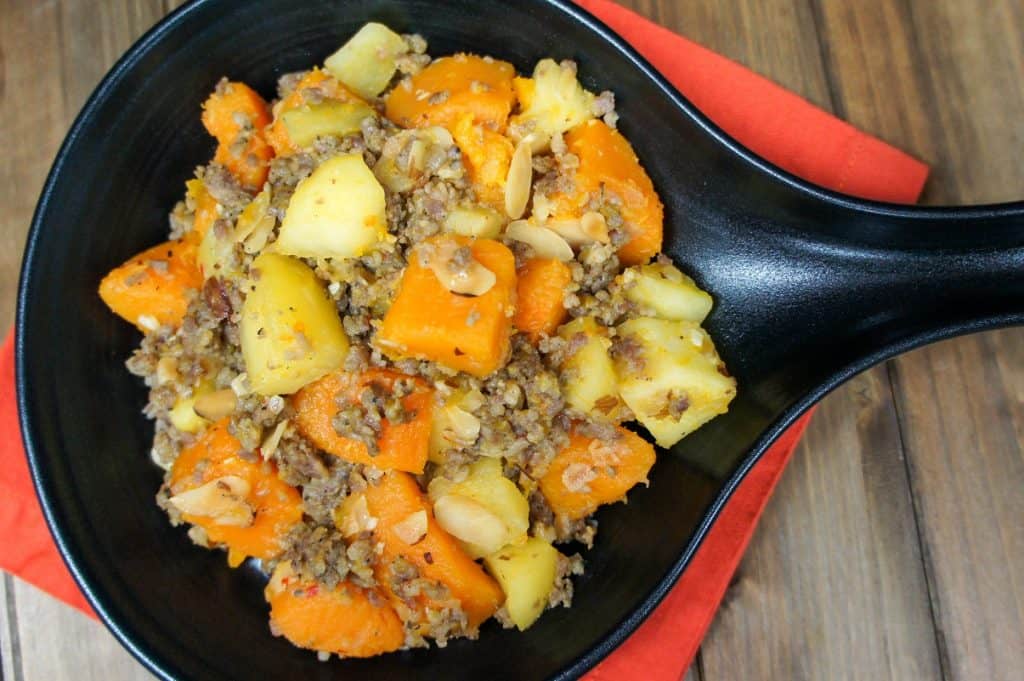 A close of the the sausage and butternut squash in a black serving skillet set on an orange linen on a wood table.