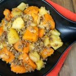 A close of the the sausage and butternut squash in a black serving skillet set on an orange linen on a wood table.