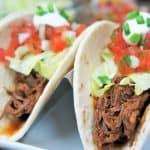 A close up of shredded beef in a soft tortilla shell topped with lettuce, diced tomatoes, sour cream and green onions.