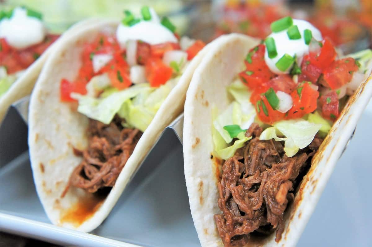 A close up of shredded beef in a soft tortilla shell topped with lettuce, diced tomatoes, sour cream and green onions.