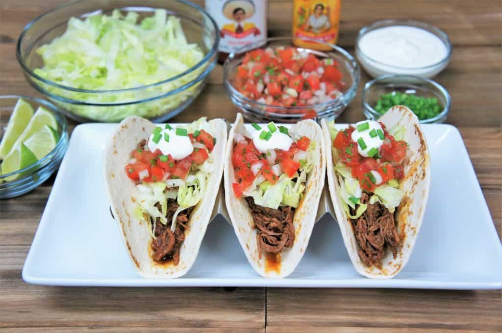 Three shredded beef tacos in a soft tortillas topped with lettuce, diced tomatoes, sour cream and green onions.