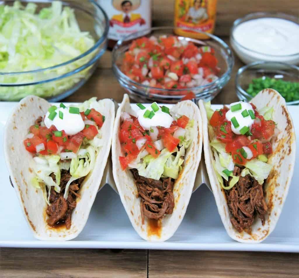 Three shredded beef tacos in soft tortillas topped with lettuce, diced tomatoes, sour cream and green onions.