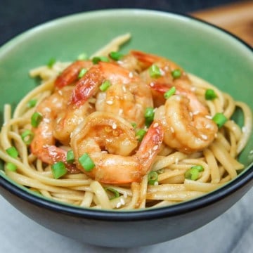 The sesame noodles topped with shrimp served in a green bowl on a beige linen on a wood table.