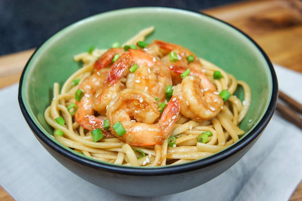 The sesame noodles topped with shrimp served in a green bowl on a beige linen on a wood table.