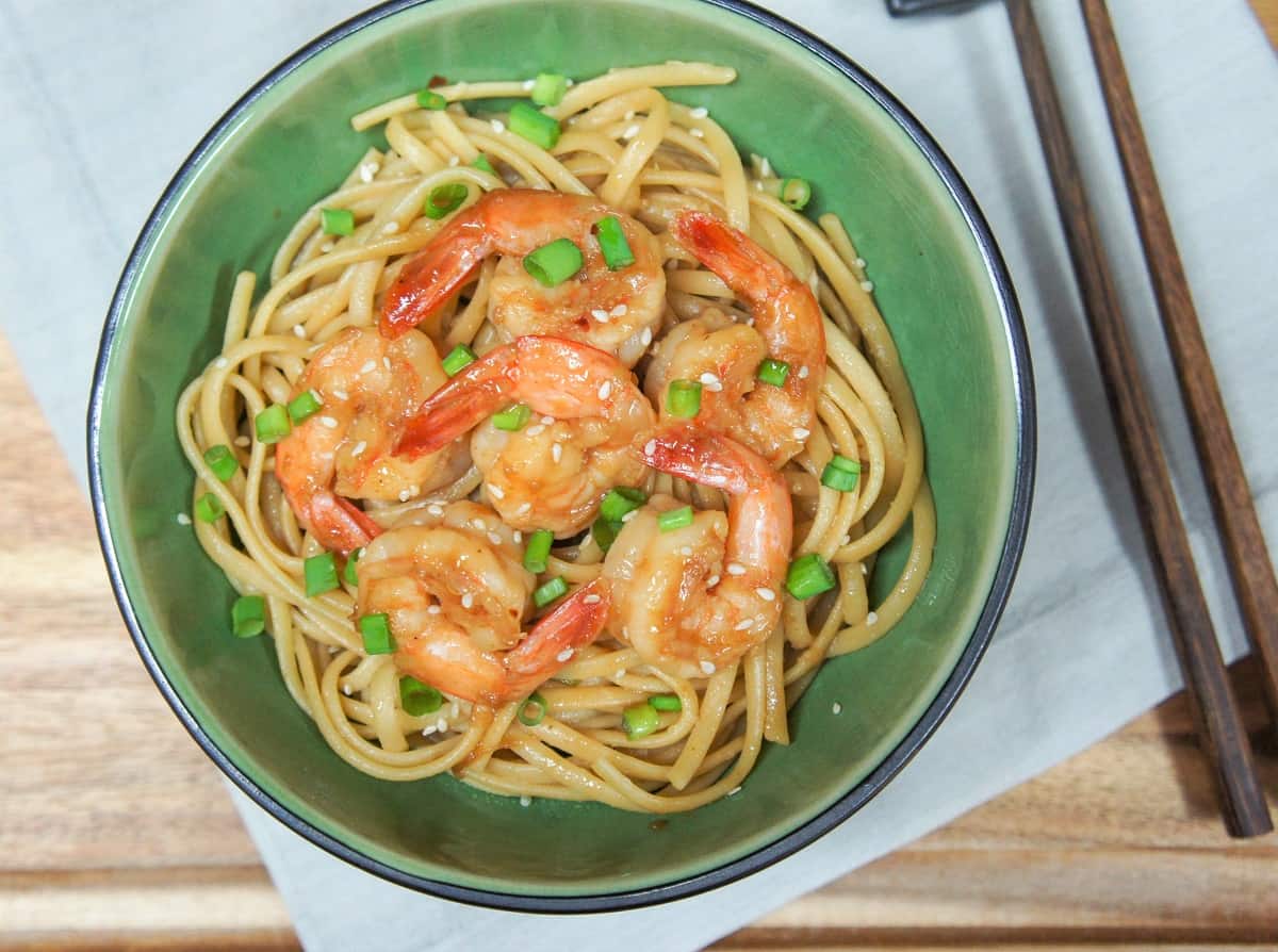 The sesame noodles topped with shrimp served in a green bowl on a beige linen on a wood table.