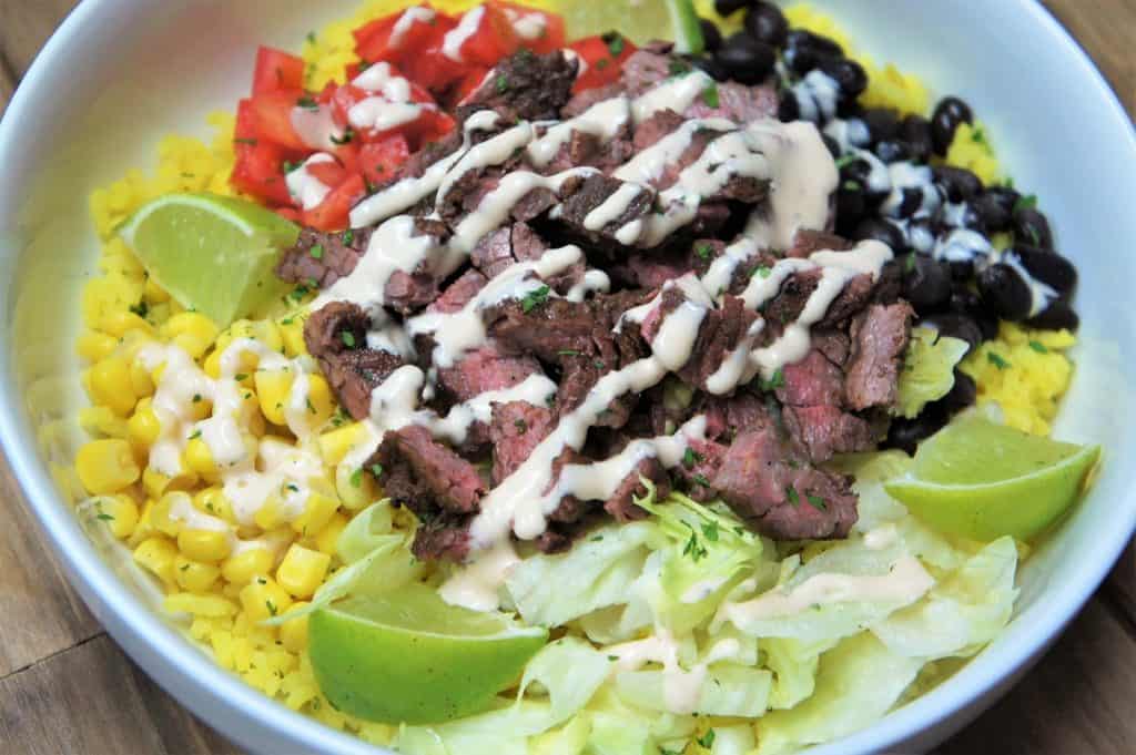 Southwestern Steak Bowl, sliced steak served on yellow rice, with black beans, corn, diced tomatoes, shredded lettuce and lime wedges all arranged on a white plate