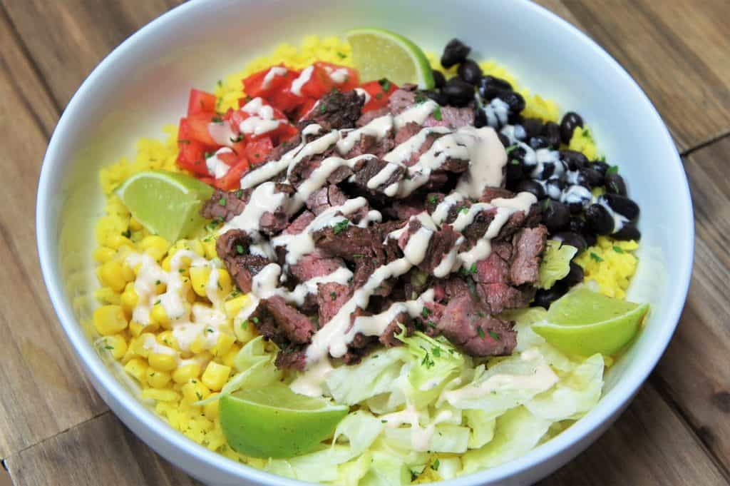 Southwestern Steak Bowl, sliced steak served on yellow rice, with black beans, corn, diced tomatoes, shredded lettuce and lime wedges all arranged on a white plate