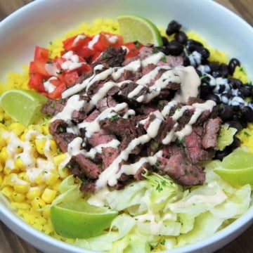 Southwestern Steak Bowl, sliced steak served on yellow rice, with black beans, corn, diced tomatoes, shredded lettuce and lime wedges all arranged on a white plate