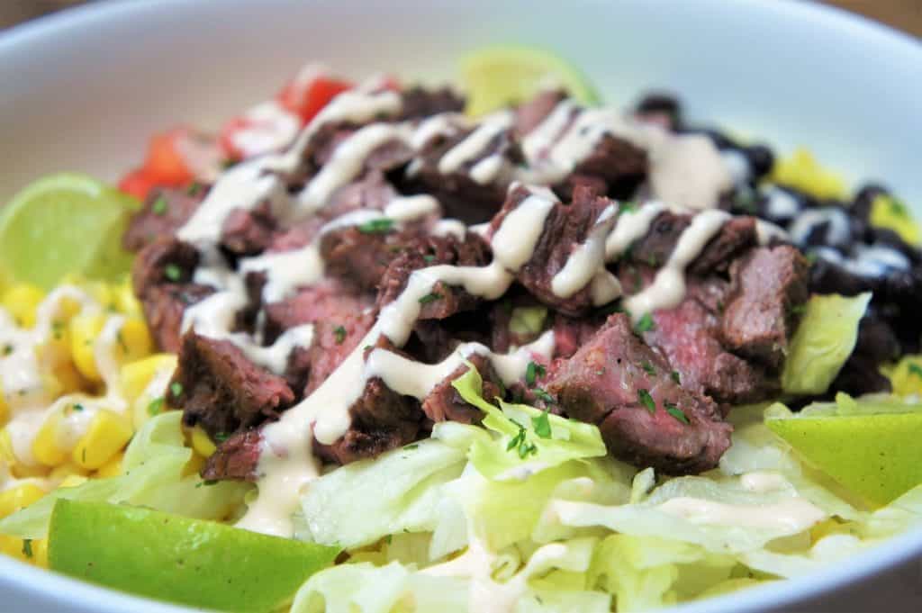 Southwestern Steak Bowl, sliced steak served on yellow rice, with black beans, corn, diced tomatoes, shredded lettuce and lime wedges all arranged on a white plate