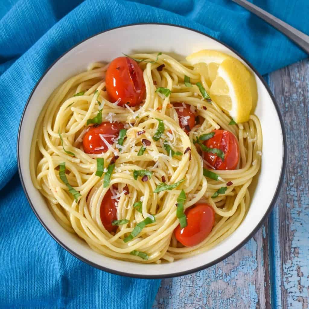 The spaghetti aglio e olio with fresh tomatoes and basil served in a white bowl with a black rim set on an aqua linen and a light blue wood table.