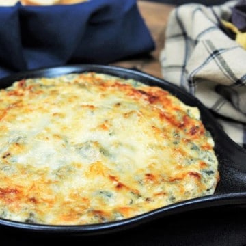 Spinach and Artichoke Dip covered with melted cheese and served in a small black skillet with corn tortilla chips and bread sliced in the background