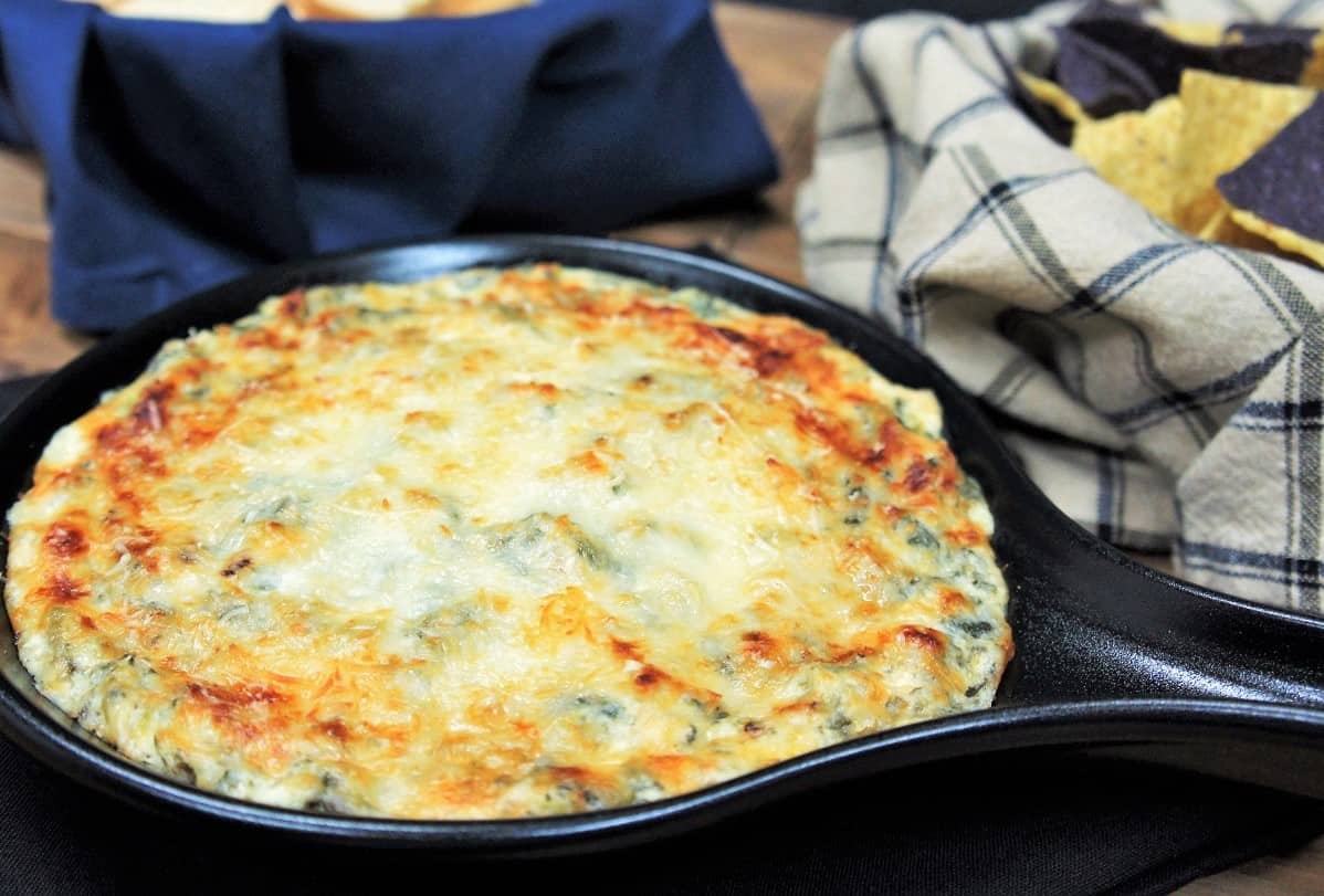 Spinach and Artichoke Dip covered with melted cheese and served in a small black skillet with corn tortilla chips and bread sliced in the background