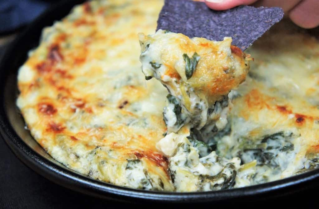 Spinach and artichoke dip served in a black skillet with some being scooped by a blue tortilla chip.