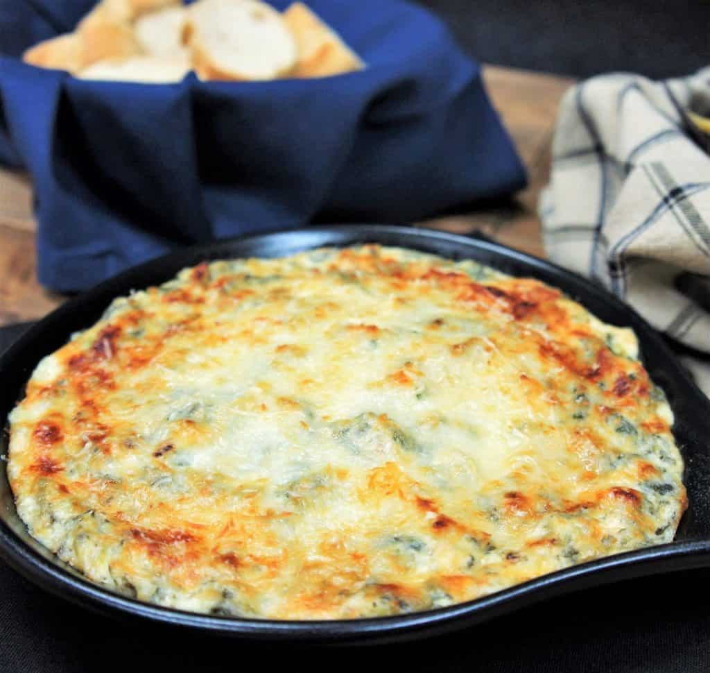 A close up image of spinach and artichoke dip served in a small black skillet with a bread basket in the background.