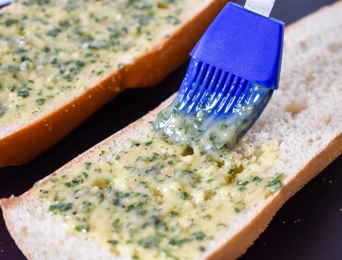 A blue pastry brush applying garlic butter to a loaf of bread that is sliced in half.