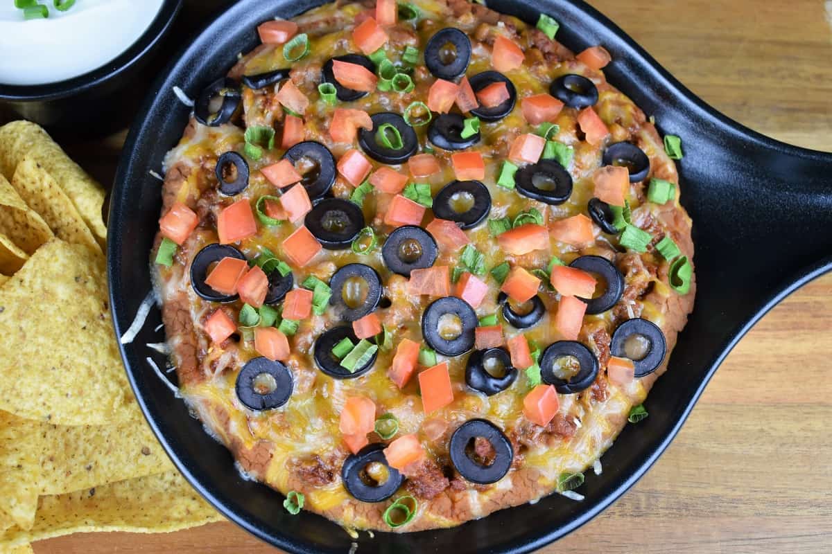 Taco Dip, refried beans and seasoned ground turkey topped with melted cheese, diced tomatoes, black olives and green onions served in a black bowl with corn tortilla chips and sour cream on the side.