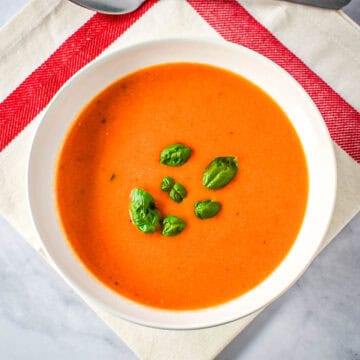Bowl of creamy tomato soup garnished with fresh basil leaves, served on a white napkin with red stripes.