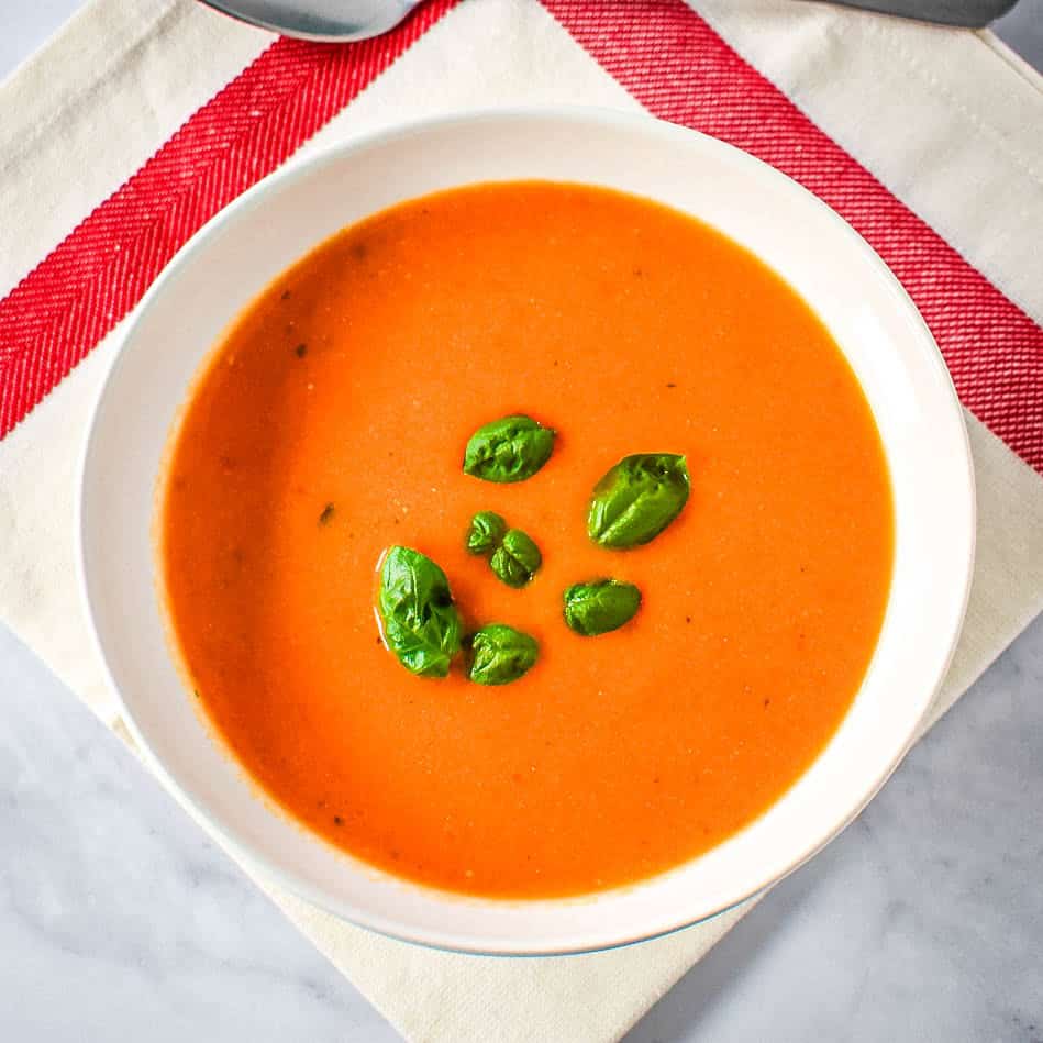 Bowl of creamy tomato soup garnished with fresh basil leaves, served on a white napkin with red stripes.