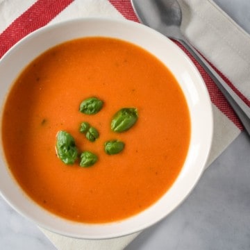 An image of the tomato soup served in a white bowl on a white linen with a red stripe and a spoon to the top, right side.
