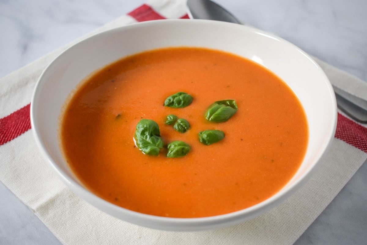 The finished soup garnished with small basil leaves and served in a white bowl set on a white linen with a red stripe.