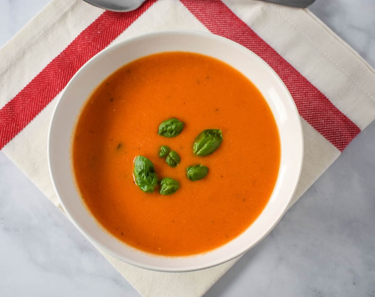 The finished soup garnished with small basil leaves and served in a white bowl set on a white linen with a red stripe.