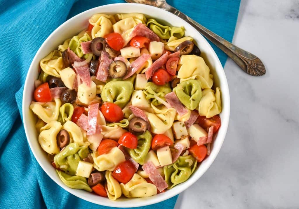 The tortellini pasta salad served in a large white bowl, displayed on a white table with an aqua colored linen underneath and a serving spoon in the background.