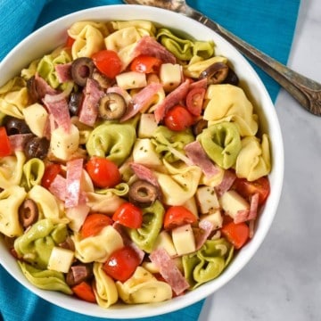 The tortellini pasta salad served in a large white bowl, displayed on a white table with an aqua colored linen underneath and a serving spoon in the background.
