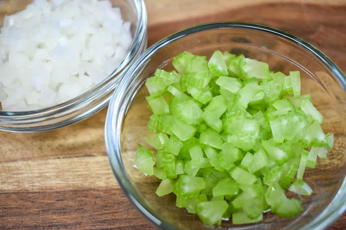Finely diced celery and onions separated in two small, clear bowls on a wood cutting board.