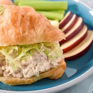 Tuna salad topped with shredded lettuce on a croissant with sliced apples and celery sticks on the side. The sandwich is served on a blue plate.