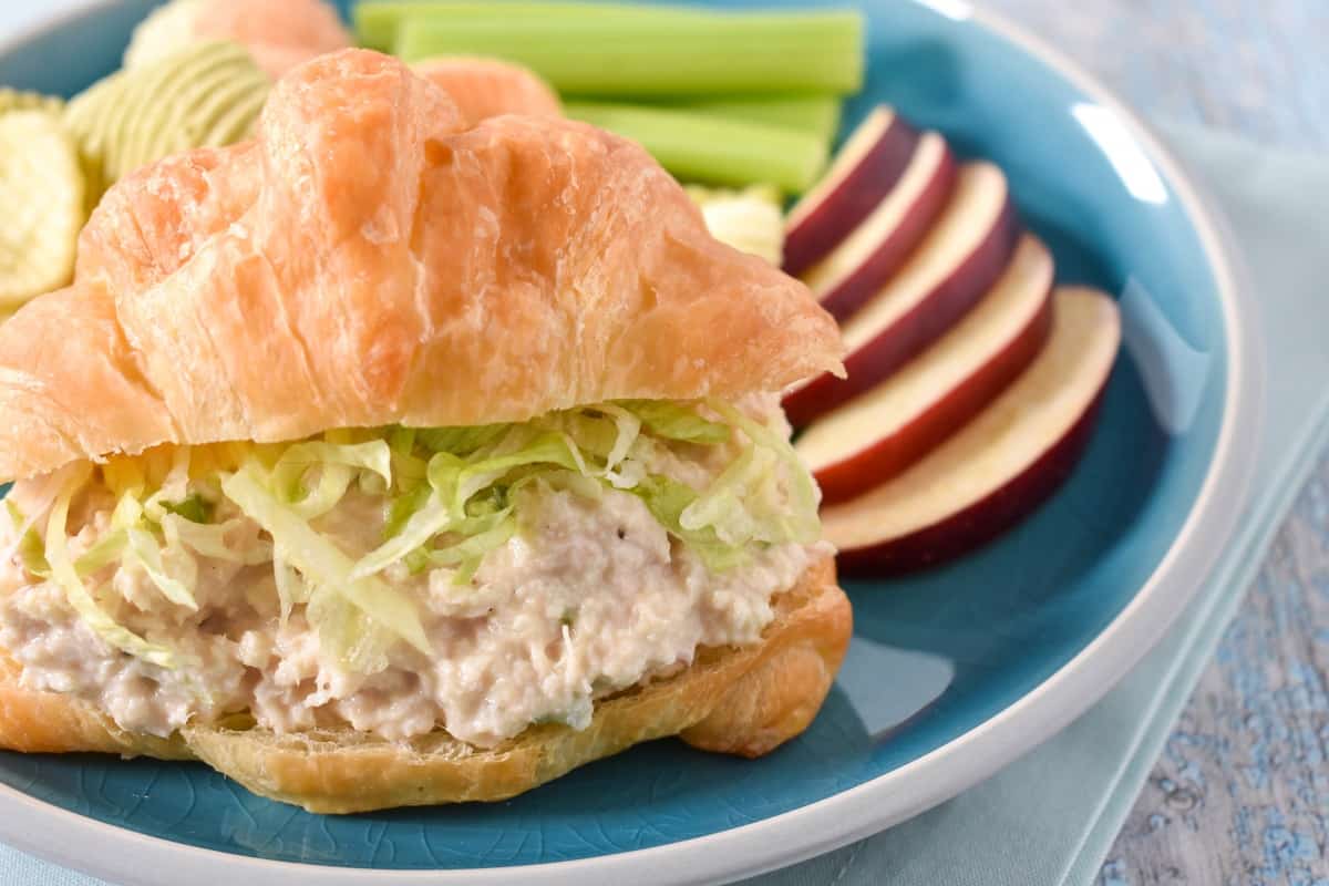 Tuna salad topped with shredded lettuce on a croissant with sliced apples and celery sticks on the side. The sandwich is served on a blue plate.