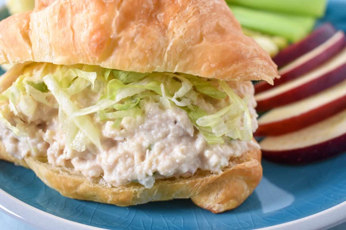 A close up image of the tuna salad served on a croissant topped with shredded lettuce with sliced apples and celery sticks on the side.