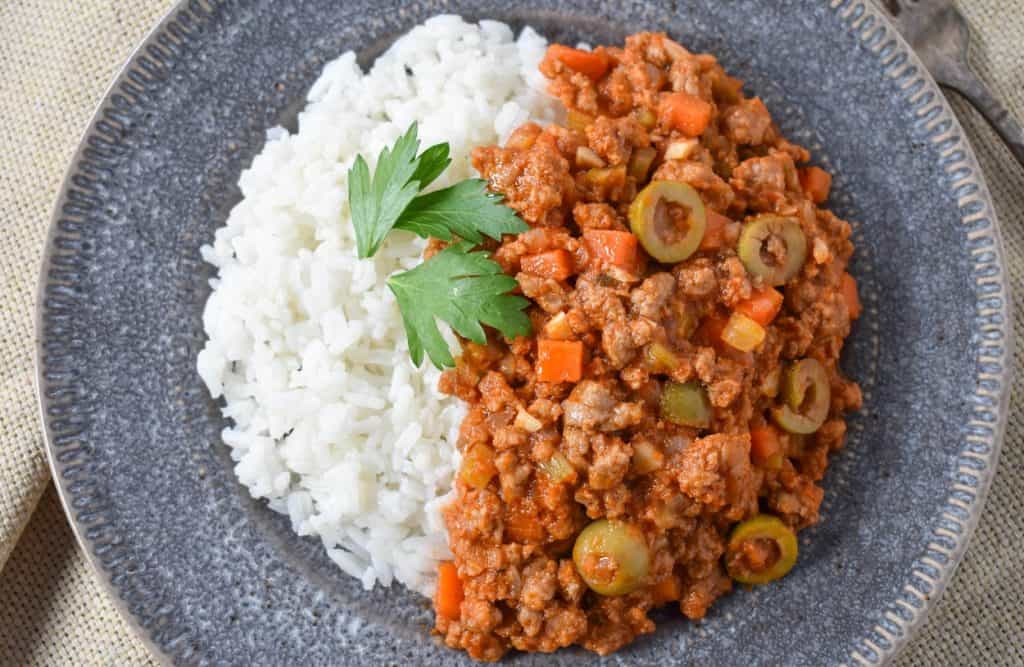 The picadillo served with white rice on the side on a gray plate and garnished with parsley leaves.