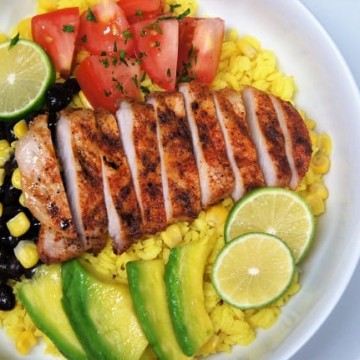 Yellow Rice + Grilled Pork Loin Bowl, a bed of yellow rice topped with grilled, seasoned pork loin, corn, black beans, sliced avocados and diced tomatoes, garnished with sliced limes