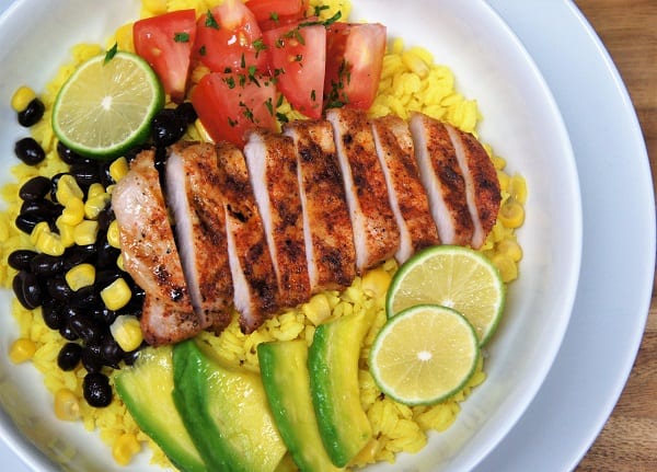 Yellow Rice + Grilled Pork Loin Bowl, a bed of yellow rice topped with grilled, seasoned pork loin, corn, black beans, sliced avocados and diced tomatoes, garnished with sliced limes