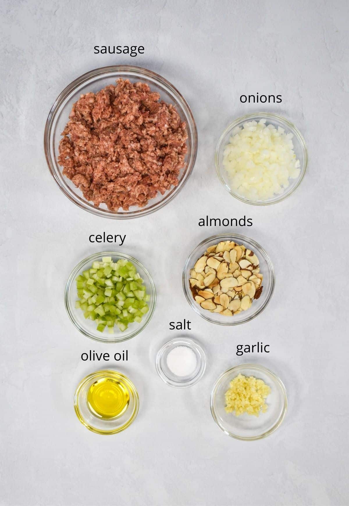The ingredient for the sausage stuffing mix prepped and arranged in glass bowls and set on a white table.