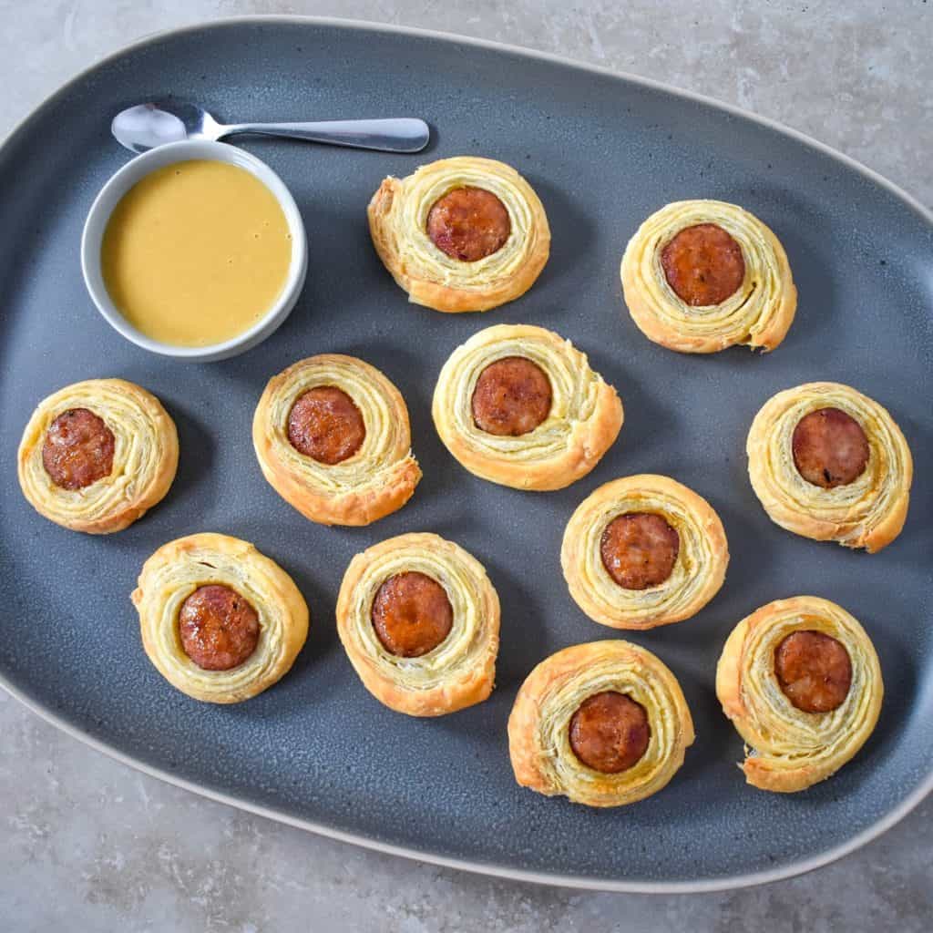 Andouille sausage puffs arranged on a large gray platter with a small bowl of the dijon honey sauce and a small spoon to the side.