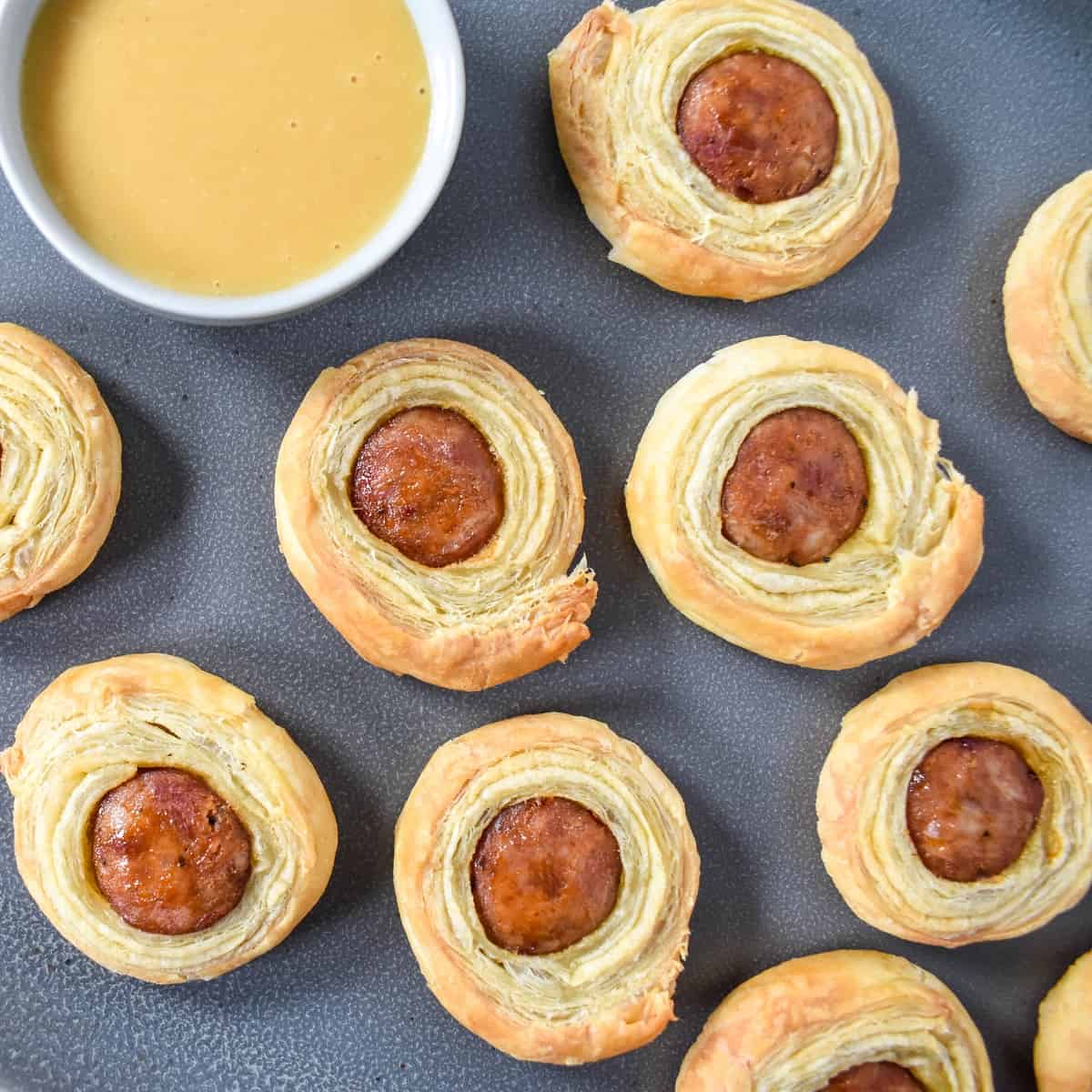 A close up image of the andouille sausage pastries arranged on a large gray platter with a small bowl of the dijon honey sauce and a small spoon to the side.