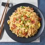 The finished bacon fried rice served on a black plate with a beige linen and chopsticks to the left side.