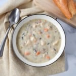 An image of the chicken and wild rice soup served in a white bowl with a black rim. The plate is set on a beige linen on a white table with a soup spoon and a piece of bread on the side.