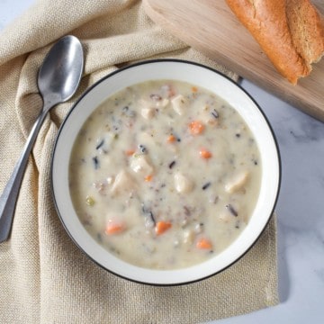 An image of the chicken and wild rice soup served in a white bowl with a black rim. The plate is set on a beige linen on a white table with a soup spoon and a piece of bread on the side.
