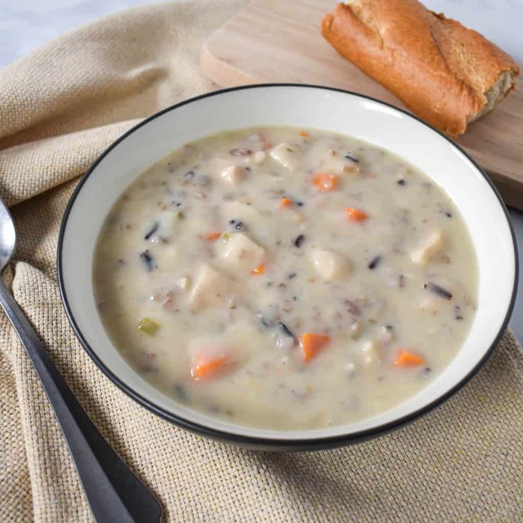 An image of the creamy chicken and wild rice soup served in a white bowl with a black rim. The plate is set on a beige linen with a soup spoon and a piece of bread on the side.