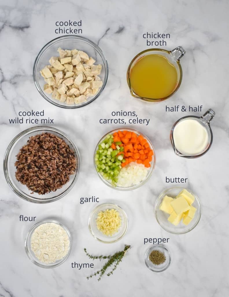 The ingredients for the chicken and wild rice soup prepped and arranged in glass bowls on a white table. Each ingredient is labeled with small letters.