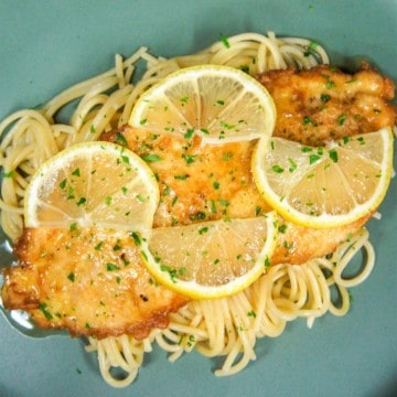 A top view image of the chicken francaise served on a bed of pasta and garnished with thin lemon slices and chopped parsley.
