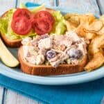 Open-face chicken salad sandwich with grapes, celery, and toasted almonds, served with lettuce, tomatoes, potato chips, and a pickle.