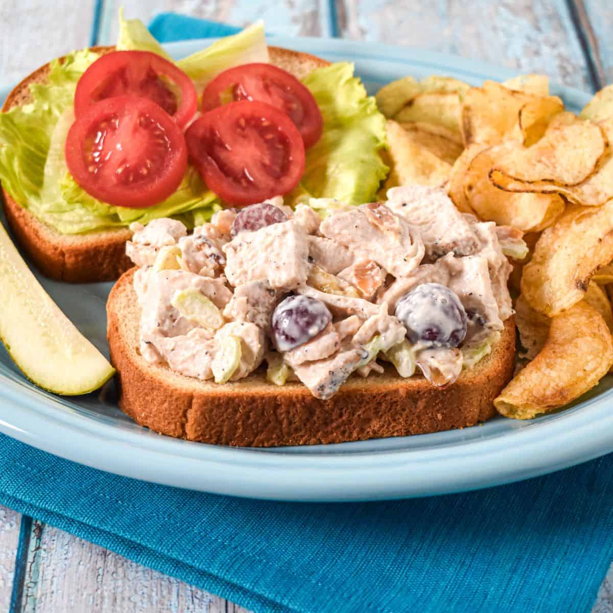 Open-face chicken salad sandwich with grapes, celery, and toasted almonds, served with lettuce, tomatoes, potato chips, and a pickle.