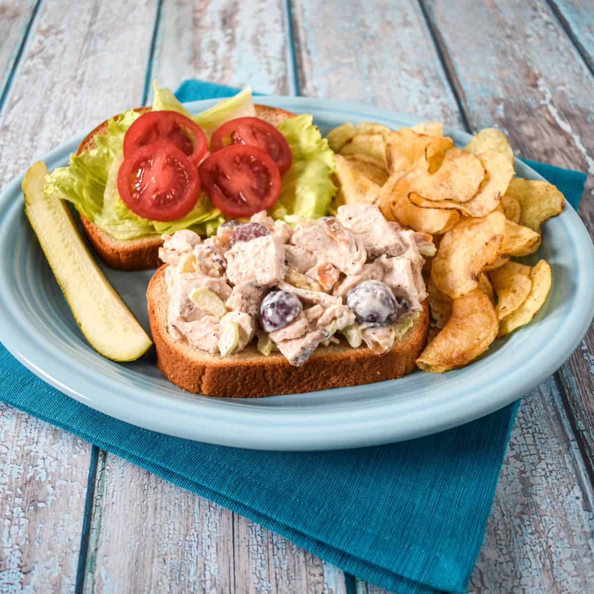 Open-face chicken salad sandwich with grapes, celery, and toasted almonds, served with lettuce, tomatoes, potato chips, and a pickle.