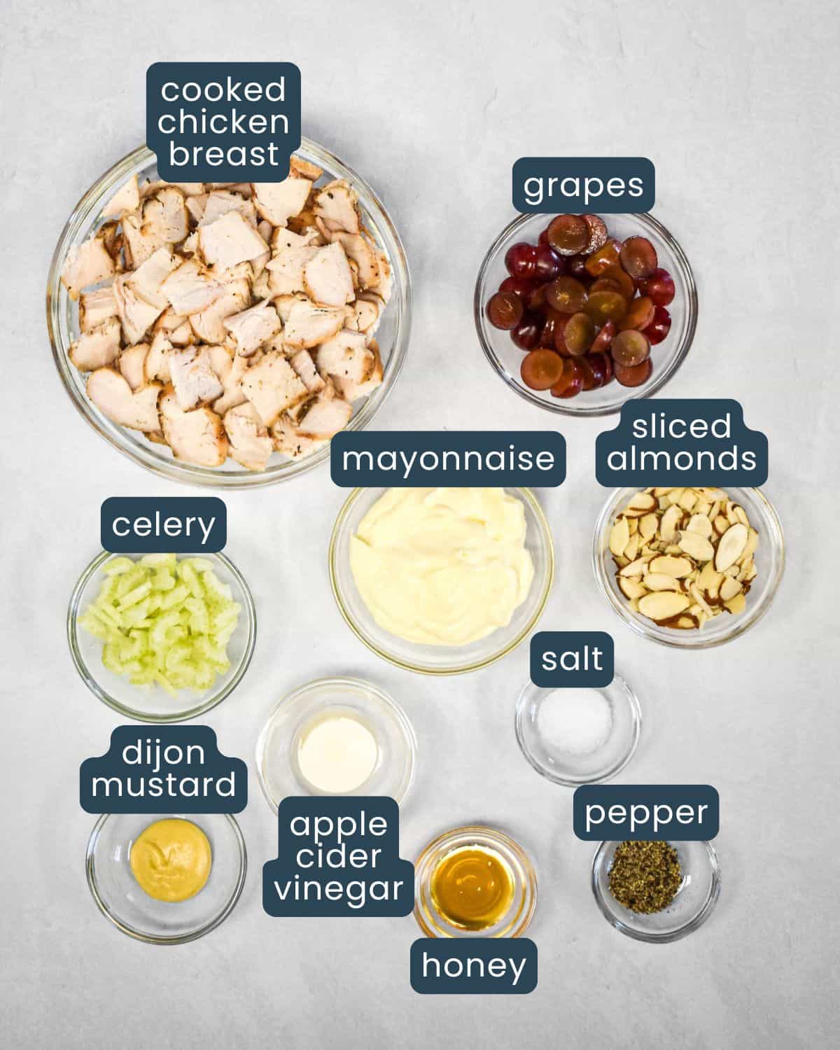 Ingredients for chicken salad sandwich including cooked chicken, grapes, celery, sliced almonds, mayonnaise, Dijon mustard, apple cider vinegar, honey, salt, and pepper, with each labeled.