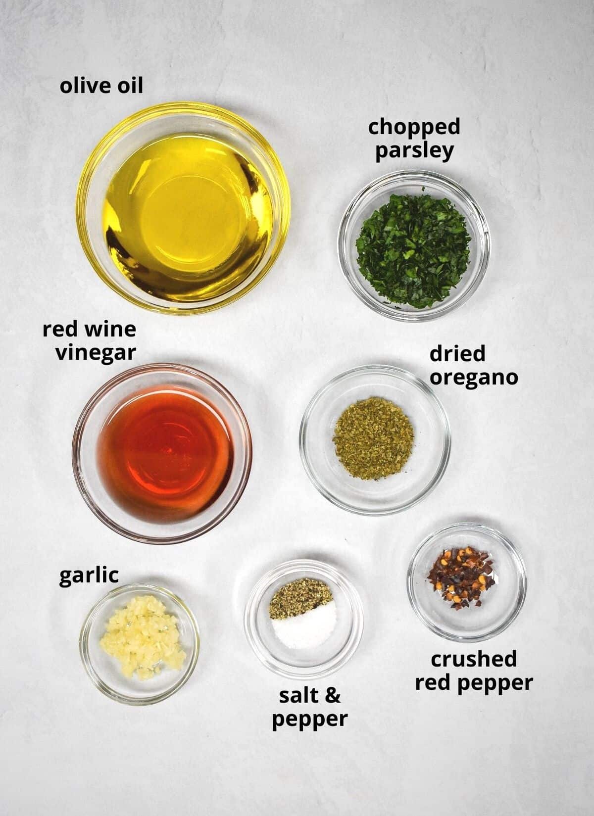 The ingredients for the chimichurri dressing arranged in glass bowls on a white table with each ingredient labeled in black letters.