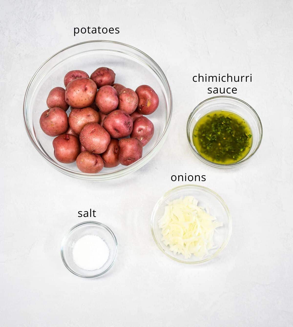 The ingredients for the potatoes arranged in glass bowls set on a white table. Each one is labeled with small, black letters.