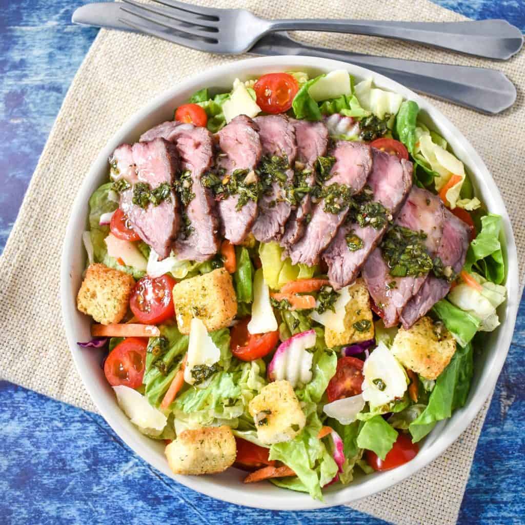 The chimichurri steak salad in a white bowl and set on a beige linen on a blue table.
